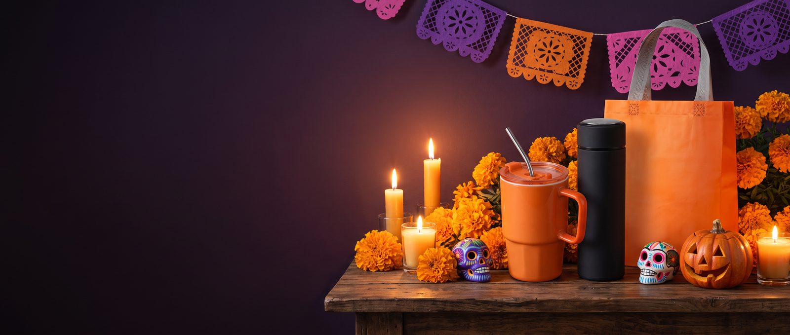 Taza naranja, termo negro y bolsa naranja en un altar con cempasúchil, calaveritas de azúcar, calabaza, velas y papel picado
